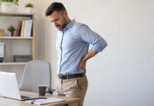 Problemas de coluna: conheça os principais tipos e como prevenir Adulto em escritório inclinado segurando lombar, ilustrando desconforto físico relacionado a problemas de coluna causados por postura inadequada.