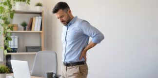 Problemas de coluna: conheça os principais tipos e como prevenir Adulto em escritório inclinado segurando lombar, ilustrando desconforto físico relacionado a problemas de coluna causados por postura inadequada.
